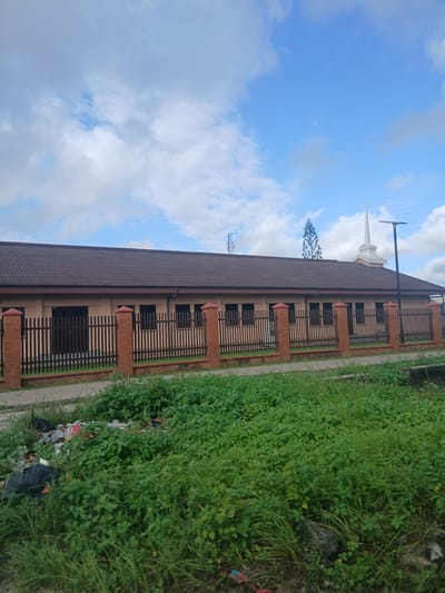 The image shows a building with a brown tiled roof and a beige facade. It is surrounded by a brick and wooden fence. A street light stands near the building. In the background, a church spire is visible, along with a tall evergreen tree. The foreground is dominated by overgrown green grass and a pile of garbage. The sky is blue with scattered clouds. The scene appears to be in a somewhat neglected urban area, possibly Satellite Town, Nigeria.