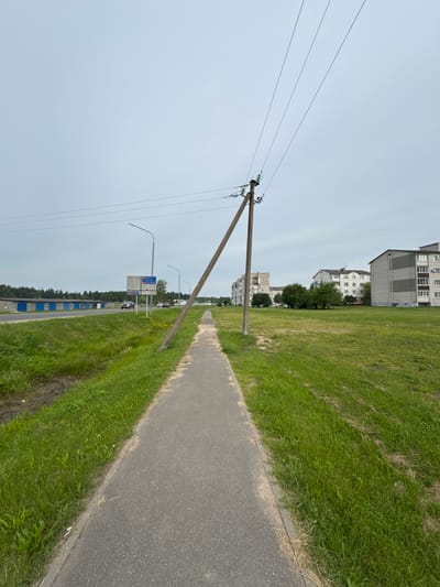 The image depicts a wide-angle view of a sidewalk in the city of Cherven, Belarus, under an overcast sky. A tilting wooden utility pole with electrical wires stretches diagonally across the frame, originating from the lower right and extending towards the upper left. The sidewalk runs parallel to a grassy median on its left, which in turn borders a road. On the far side of the road, a row of blue-doored garages is visible, followed by a treeline. Further in the background, several multi-story apartment buildings, characteristic of Soviet-era architecture, are scattered across the landscape. The scene is quiet, with no people or vehicles immediately apparent in the foreground. The overall impression is one of a residential area on a cloudy day.