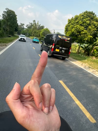 The image presents a tilted, eye-level view, focusing on a human hand in the immediate foreground, with the middle finger extended outward. This gesture appears directed towards a road scene in the background, conveying defiance or insult.
The setting is an asphalt road in Vị Xuyên Commune, Vietnam, clearly marked with a solid yellow line down its center and a white line along its right shoulder. Several vehicles are visible on the road. Most prominent is a black passenger van, possibly a commercial vehicle, with the word "LUXURY" clearly visible on its rear. Below "LUXURY", a Vietnamese license plate appears to display "30A-000.35". Further down the road, a turquoise car and a white car can be seen.
The road is bordered by dense, lush green trees and tropical foliage on both sides, indicative of a verdant rural or semi-rural landscape. The sky above is clear with scattered clouds, suggesting a bright daytime, likely late morning or early afternoon, with ample natural light.