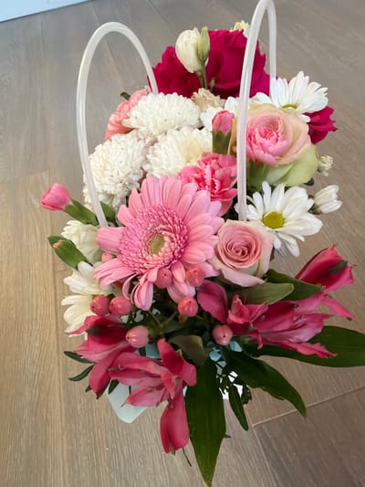 A high-angle shot shows a bouquet of flowers in a white basket with a clear plastic handle, placed on a light-colored wooden floor. The bouquet contains various flowers in shades of pink, white, and dark red, including roses, gerberas, carnations, daisies, and alstroemerias, as well as small pink berries. The background is a blurred light blue wall. The setting is indoors in the city of Uzyukovo, Russia.