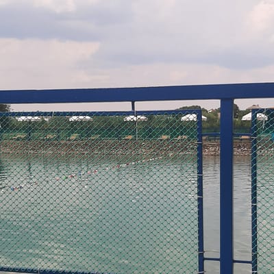The image depicts an outdoor scene in Dubrivka, Ukraine, likely a recreational area. In the foreground, a blue metal railing runs horizontally across the top of the frame, with vertical blue posts supporting it. A green mesh fence is visible in front of the railing, obscuring part of the view.
Beyond the fence, a body of water stretches into the distance, appearing to be a pond or small lake. Scattered across the water's surface are numerous small, brightly colored buoys, some red, white, blue, and green, indicating they are likely marking a course or area. The water is calm with subtle ripples.
In the mid-ground, on the far bank of the water, a line of white umbrella-like structures can be seen, suggesting some sort of seating or shelter area. Behind these, trees form a dense green backdrop, with glimpses of rooftops of buildings peeking through the foliage, indicating the presence of a settlement. The sky above is overcast with a mix of white and gray clouds. The overall impression is a calm, possibly a fishing or boating facility.