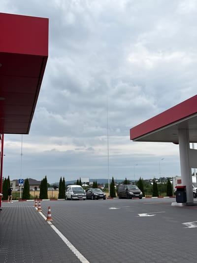 This image shows a view of a gas station under a cloudy sky. In the foreground, a paved lot with red and white striped borders is visible. Several orange traffic cones are placed on the lot. To the left, a section of a red-roofed structure extends into the frame. In the mid-ground, a white van and a dark-colored sedan are parked on the lot. Further back, a dark brown van is also parked. A person in dark clothing and shorts is walking on the paved area to the left. Behind the parked vehicles, a row of tall, slender green trees is lined up, with buildings and houses visible in the distance. The overall environment appears to be a commercial area within a town or city.