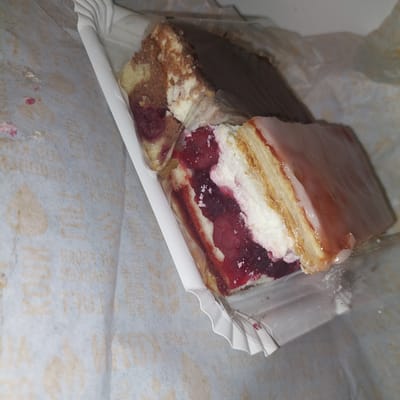 A close-up, overhead shot captures two slices of cake within a white paper tray. The cake on the left is a marbled cake with layers of chocolate and vanilla cake, interspersed with cream and a dark red fruit filling, possibly cherries. It is partially covered by a clear plastic wrap. The cake on the right is layered, with a creamy white filling between thin cake layers, and topped with a pale pink glaze. The tray is placed on a surface covered with patterned paper, which appears to be light gray with a subtle, repeated beige pattern. The overall scene suggests a close-up of desserts, likely taken indoors. There is no indication of a specific location in Landshut, Germany.