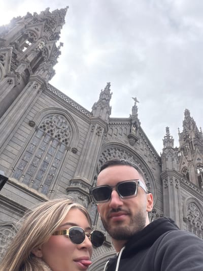 Two individuals are depicted in a selfie taken in front of the Church of San Juan Bautista in Arucas, Gran Canaria, Spain. The male subject, positioned on the right, has short dark hair and a beard, wears square grey-framed sunglasses, and a dark hooded sweatshirt. He looks directly at the camera. The female subject, positioned on the left, has long blonde hair with highlights, wears round gold-framed sunglasses, and is shown with her tongue extended playfully. Both individuals exhibit light skin tones and appear to be in their 20s or 30s. The background is dominated by the monumental Neo-Gothic architecture of the church, constructed from dark volcanic stone, featuring a tall, ornate spire, pointed arches, intricate tracery, and stained-glass windows. The sky overhead is overcast and grey.