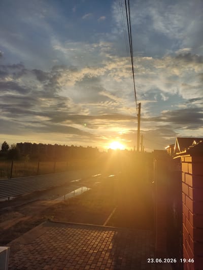 This image depicts a sunset over a landscape in Kuyuki, Russia. The sun is a brilliant orb, low on the horizon, casting a warm, golden glow across the scene. Wispy clouds, tinged with yellow and orange, stretch across the sky, creating a dramatic backdrop for the sunset.

In the foreground, a power line stretches horizontally across the frame, supported by a utility pole on the left. To the right, a brick wall forms a vertical element, its textured surface catching the warm light. Beyond the power line and to the right, a silhouetted cityscape with numerous buildings is visible against the bright sky. The bottom right of the image shows a brick sidewalk or pathway, adding to the urban or semi-urban context of the scene. The overall mood is serene and peaceful, evoking the quiet beauty of a day's end.

The visible text in the image indicates the date and time: "23.06.2026 19:46", confirming it was taken in the late evening on June 23, 2026. The weather appears clear, with the clouds adding visual interest to the sunset.