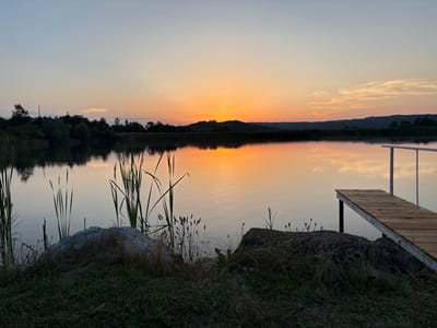 This is a photograph of a calm lake at sunset, likely located near Skalsko, Bulgaria. The sky displays a gradient of colors, transitioning from a pale blue at the top to a vibrant orange near the horizon where the sun is setting behind a dark, silhouetted hill. The warm hues of the sunset are reflected beautifully on the still surface of the water.
In the foreground, there is a wooden dock extending into the lake from the right side of the frame. The dock is constructed of weathered wooden planks and has white metal supports. On the left side of the lake's edge, large rocks and tall reeds or cattails are visible, with green grass growing around them. The reflection of the reeds and rocks can be seen in the water. The overall atmosphere is serene and tranquil, capturing the peaceful end of a day. There are no people or animals visible in the image.