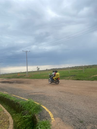 The image depicts three individuals on a motorcycle on a paved road. The person in the front is wearing a dark shirt, while the passenger behind them is in a yellow shirt. The third person, riding on the back, is wearing a dark shirt and appears to be leaning forward. They are traveling down a road that has a yellow line marking its edge in some sections and grass growing along the sides. Beyond the road, there is a vast, green, open field with scattered trees and what appear to be fence posts. In the distance, the sky is overcast and appears heavy with clouds, suggesting it might be a cloudy day. The overall scene has a rural or semi-rural feel, possibly in a developing area given the motorcycle and the open fields.
The image is oriented sideways, which might suggest it was taken while the motorcycle was in motion or during a casual stop. The lighting is diffused, consistent with an overcast sky, and there is no strong indication of the time of day beyond it being daytime. The location is identified as Bulamare, Nigeria. There are no visible text elements within the image.