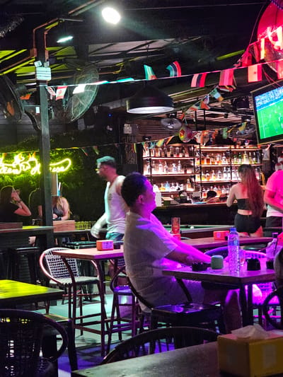 The image depicts a dimly lit, outdoor bar or restaurant scene in Ban Klong Haeng, Thailand, likely at night. The setting is cluttered and casual, with various objects visible, including shelves stocked with bottles of alcohol behind a bar, several fans, chairs, and what appear to be national flags strung up as decorations. A large television screen displaying a sports game is mounted above the bar.

There are several people present, though their activities are somewhat obscured. In the foreground, a man in a white shirt is seated and appears to be interacting with someone off-camera or looking down. Further back, a woman with long, curly hair is visible near the bar, and another person is seated and looking up at the camera. The overall atmosphere seems relaxed and social, consistent with a bar setting.

Notable details include the neon yellow "Chiang Mai" sign, a clear indicator of the location. The lighting is a mix of ambient bar lights, neon signs, and some spotlights, creating a vibrant yet somewhat shadowed environment. The presence of multiple fans suggests a warm climate, typical of Thailand. The image is oriented vertically, which might be a stylistic choice or a result of how the photo was taken. The image is a photograph.