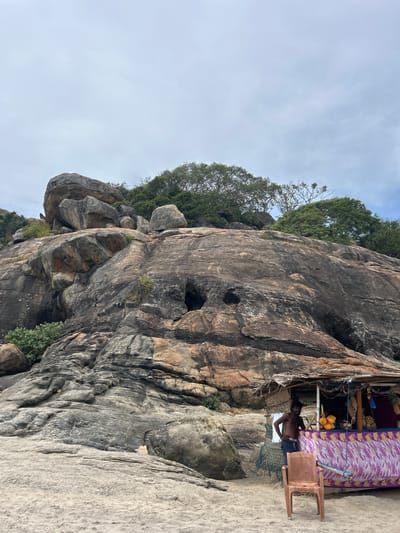 This is a ground-level, vertical, eye-level, medium shot photograph of a beach scene in Shastrawela, Sri Lanka. In the foreground, a sandy beach transitions to a large rock formation that dominates the midground and background. The rock face is textured with varying shades of gray, brown, and orange, and features several dark crevices and openings. Trees with lush green foliage grow from the top of the rock formation.

On the right side of the image, a small roadside stall is set up against the rock face. The stall has a thatched roof and is decorated with colorful fabrics and festive garlands. A shirtless man with dark hair and a light-colored waistband is standing behind the counter of the stall, looking towards the camera. He appears to be the vendor. In front of the stall, a single, empty, brown plastic chair sits on the sand. The stall displays an assortment of fruits, including pineapples and oranges. The sky above is overcast with light gray clouds.