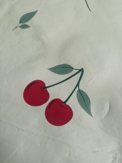 This is a close-up, top-down shot of a light-colored fabric with a printed cherry pattern. The main subjects are two red cherries with green stems and leaves, depicted in a stylized, graphic manner. One cherry is slightly larger and positioned to the left, while the second, smaller cherry is below and to the right of the first. Both cherries have a darker curved line indicating a shadow or indentation. To the upper left of the cherries, there is a sprig with two green leaves. Another single green leaf is visible to the right of the cherries, and a stem fragment is present in the upper center of the image. The fabric appears to be somewhat textured and creased, suggesting it might be a tablecloth, a piece of clothing, or bedding. There are no people or animals visible, and the scene is a simple, flat surface with a decorative print. The lighting is even and diffused, providing no strong shadows that would indicate a specific time of day or weather conditions. There is no visible text within the image. The location context of Grudziądz, Poland, does not provide any specific visual cues within the image itself.