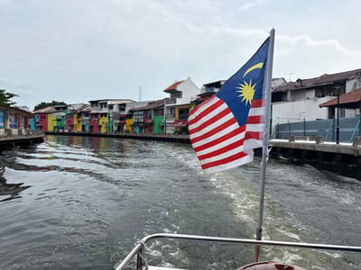 A view from a boat traveling down a canal. Colorful buildings line the canal's edge. A Malaysian flag is prominently displayed on the boat, partially obscuring the view. The water reflects the overcast sky. The boat's wake trails behind, indicating its movement.