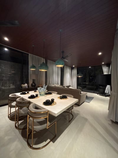This image displays a modern dining and living area within a property located in Batuan, Indonesia. The interior is artificially lit, suggesting an evening or nighttime setting due to the dark outdoor environment visible through extensive glass panels.

In the foreground and midground, a rectangular white marble dining table is centrally positioned. Eight contemporary dining chairs, featuring gray upholstered seats and backs with gold-toned metal frames, are arranged around the table. Each place setting on the table consists of a small gold-colored placemat, a black saucer, and a black cup. A large, decorative glass vase with swirling blue and white patterns serves as the centerpiece.

The ceiling is clad in dark red-brown slatted panels, from which four teal-colored pendant lights, a dark ceiling fan, and multiple recessed spotlights are suspended, providing illumination. The walls on the left and far side of the room are predominantly floor-to-ceiling glass windows, draped with light-colored vertical curtains that are partially drawn open. A dark, possibly brick or stone, wall section is visible on the far left.

To the right of the dining area, a light-toned sofa with multiple cushions defines a living space. A blue rectangular rug is placed on the floor in front of the sofa. A large, dark flat-screen television on a stand is situated towards the far right. The floor throughout the visible area consists of large, light-colored tiles. A dark console table holding a lamp with a black shade and a woven basket is positioned along the far left wall. No individuals are present in the scene.