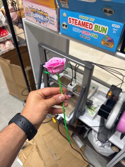 A person's hand, wearing a black smartwatch, holds a pink 3D-printed rose with a green stem. The hand is positioned in the foreground, with the rose extending upwards and slightly to the left. In the background, there are shelves stocked with various items, including toy boxes with colorful packaging. One box is bright blue and features text that reads "SQUISHY TOY STEAMED BUN SOFT CUTE FUN DO NOT EAT." Another box, partially visible, is pink and has text that includes "GOOD DUMPLING SURPRISE." Behind the shelves, a gray 3D printer is visible with its mechanical components and spools of filament. The environment appears to be an indoor retail or workshop space. The image was taken in Keila, Estonia.
