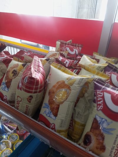 The image depicts a retail display of numerous individually packaged croissants inside a store in Naung Bo Oe, Myanmar. The pastries are primarily branded as "5 DAYS Croissant," with several packages featuring a "HALAL" certification logo. Packaging designs vary, including red and white plaid patterns, and cream-colored wraps with images of croissants showing different fillings such as yellow cream and a dark berry-like substance. The packages are stacked upright in multiple rows within a display unit, which has a metal railing in the foreground. Below this display, part of a blue bin is visible, containing several small, round containers with yellow lids, possibly yogurt or similar products. The background features a prominent solid red panel extending horizontally, with a blurred view of an exterior or another section of the store visible above it, showing vertical light-colored structures and ambient light. No individuals or explicit actions are visible within the frame.