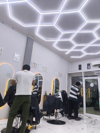 This image captures the interior of a modern barbershop in Miruektaman, Indonesia. The dominant feature of the ceiling is a pattern of interconnected hexagonal LED light fixtures emitting bright white light. The walls are painted white, with a gray rectangular column on the left side. Along the left wall, several arched mirrors are mounted, each illuminated from behind with a warm orange-yellow glow, and framed certificates or documents hang between them.
In the foreground, a man wearing a white short-sleeved t-shirt and olive green trousers stands with his back to the camera, attending to a client seated in a black hydraulic barber chair. The client is mostly obscured by the barber and a black cape. Further to the right, another male barber, wearing a horizontally striped t-shirt (dark and white) with "GOOD -ATTITUDE-" printed on the back and dark trousers, is also seen from behind, actively cutting a client's hair with clippers. The client, a young man with dark hair draped in a black cape, is visible in the reflection of a mirror behind the second barber.
The floor is covered with white tiles and is visibly strewn with numerous dark hair trimmings, particularly around the barber chairs, indicating active business. On the right side of the room, a white split-unit air conditioner is mounted high on the wall. A glass double door with a white frame leads to an adjacent area. A circular white sticker on the right door panel features the word "CLOSE" in black and a stylized graphic, with "SUHU BARBER" written vertically below it. Through this glass door, a person wearing green clothing is faintly visible seated inside.