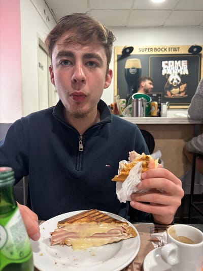 This image depicts a young male seated at a table within a casual dining establishment in Gondomar, Portugal. The subject, with short brown hair, blue eyes, and light facial hair, is looking directly at the camera with his mouth slightly open. He is wearing a navy blue quarter-zip fleece top with a small logo on the zipper pull. In his right hand, he holds a partially consumed grilled ham and cheese sandwich, wrapped in white paper. On the white plate before him rests another half of a grilled ham and cheese sandwich, exhibiting melted cheese and distinct grill marks on the bread. A green beverage bottle is partially visible on the left side of the frame, and a white espresso cup with a saucer, containing coffee residue, is positioned on the right. The table is covered by a brown paper placemat with faint printing. The background reveals a counter area featuring a large dark sign. The sign displays "SUPER BOCK STOUT" at the top, an illustration of a dark beer glass on the left, and a cartoon panda eating pizza with text "COME FOME PANDA" on the right. A bearded male is visible behind the counter, looking to the right. Another patron's arm and torso, wearing a grey patterned garment, are partially seen on the far right. Various condiment bottles and metal containers are present on the counter. The interior walls and ceiling are white, with a pinkish light illuminating a section of the left wall.