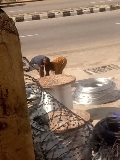The image depicts an outdoor, sunlit scene on an unpaved roadside in Arun, Nigeria. A large, cracked, brown vertical structure occupies the extreme left foreground. In front of this, and extending right, are multiple stacks of concertina razor wire, with some resting on large wooden cable spools. Another large, empty wooden cable spool is visible on the dusty ground further right. Adjacent to it, a large, coiled bundle of bright silver-colored wire is present. To the bottom right, dark coils of electrical cable are visible.
In the mid-ground, a person wearing a dark-colored short-sleeved shirt and dark trousers is bent at the waist, facing away from the camera, seemingly attending to something on the ground. A bicycle wheel is partially visible behind this individual. Next to the person, a brownish-orange animal, consistent with a woolly sheep or goat, stands facing away from the camera. A metal grate is embedded in the ground to the right of these figures.
The background features a paved two-lane road with a solid white shoulder line and a dashed white center line. A black and white striped concrete curb separates the road from the unpaved area of the scene.