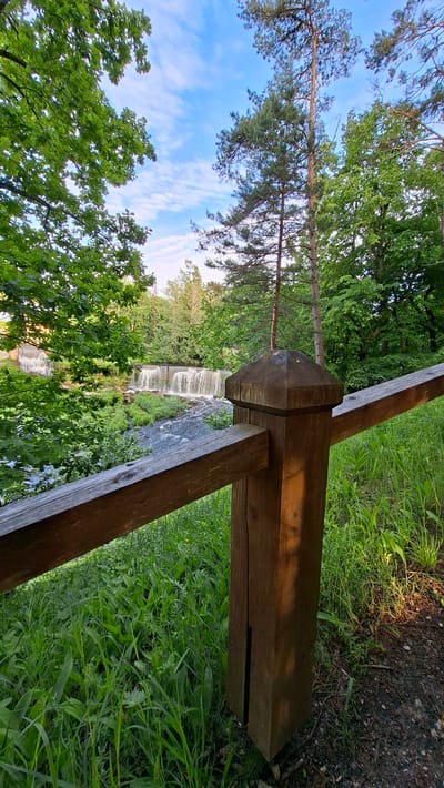 The image depicts a scenic view of the Keila-Joa waterfall in Keila-Joa alevik, Estonia. In the foreground, a weathered wooden fence post with a multi-faceted top supports a horizontal wooden railing that extends diagonally across the frame. Lush green grass and vegetation cover the ground around the fence.

Beyond the fence, a river flows, leading to a picturesque waterfall cascading over a series of rocky ledges. The water is a deep blue-grey, with white foam indicating the falls. Surrounding the river and waterfall is dense, verdant foliage, with trees and bushes forming a natural backdrop. In the distance, taller trees, including pine trees, rise towards a bright blue sky dotted with white, wispy clouds. The overall impression is one of natural beauty and tranquility.