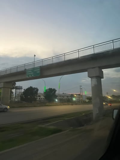 The image shows an overpass in Ade, Nigeria, viewed from a car. The overpass is made of concrete and has a railing on the side. There are streetlights with green lights on the side of the road. In the background, there are buildings and trees. The sky is cloudy, and it appears to be dusk. There are cars on the road. A green sign with white text is visible on the underside of the overpass, but the text is not legible. The overall impression is that of a typical urban scene in Nigeria during twilight.