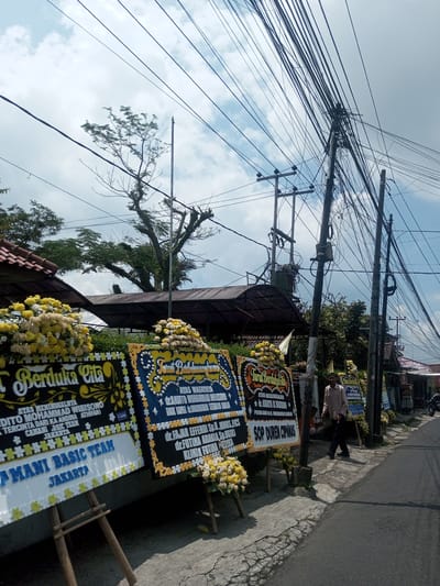 The image captures a street scene with several condolence wreaths lined along the sidewalk. The wreaths are decorated with yellow and white flowers and have inscriptions in what appears to be Indonesian. They convey condolences for the passing of "Dito Mohammad Wibisono." A man is walking past the wreaths on the sidewalk. In the background, buildings and trees are visible. Overhead, a dense network of power lines crisscrosses the sky. The location is unidentified as Xayabury District, Laos.