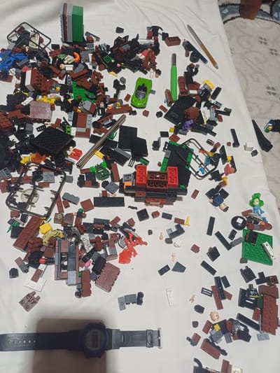 The image depicts a chaotic scattering of Lego bricks and other small toy components spread across a white surface, likely a bedsheet or tablecloth. The items are predominantly red, brown, black, and green, with some grey, blue, and yellow pieces interspersed. Among the Lego pieces are a green toy car, a disassembled plastic frame, a stylus, two pens, a watch with a digital display showing "5:30", and several small toy figures. One figure is a woman with green hair wearing a blue dress, and another is a man with a beige head. The overall impression is one of a disassembled construction project or a collection of toys in disarray. The background shows a patterned rug, suggesting an indoor setting. There is no indication of the city of Jumashuy, Uzbekistan, or any elements that would place the scene there.