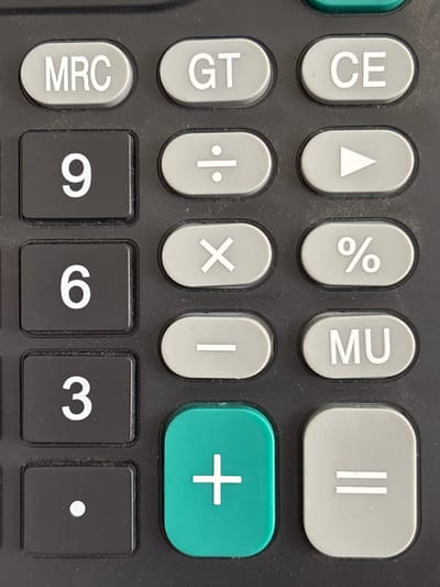This is a close-up, eye-level shot of a calculator's keypad. The calculator is black with light gray and green buttons. The buttons visible include MRC, GT, CE, division, a play symbol, percentage, multiplication, subtraction, MU, the numbers 9, 6, and 3, a decimal point, and the addition and equals symbols. The calculator is dusty, suggesting it is not in a pristine environment. The background is dark and out of focus, providing no additional environmental context. The image is focused on the central portion of the keypad. There are no people or any other elements visible in the image. There is no information to determine its location in Jrarat, Armenia.