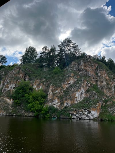 An overhanging cliff face rises steeply from a dark, rippling body of water. The cliff is a mix of grey and orange rock, with patches of green vegetation clinging to its surfaces. Trees, including pines and deciduous trees, are densely clustered on the summit of the cliff. At the very top, a small, domed structure with arched openings is visible, partially obscured by trees. The sky above is dramatic, with large, grey clouds partially obscuring the sun, which creates bright shafts of light. The scene is likely a natural landscape with a man-made structure on the cliff, possibly in the vicinity of Kamensk-Uralsky, Russia, given the context.