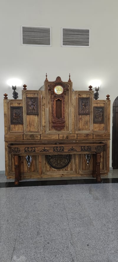 The image depicts a large, ornate wooden cabinet with a central clock and carved panels, set against a plain white wall. Two wall-mounted sconces with decorative metalwork are visible on either side of the cabinet, and two ventilation grilles are mounted higher on the wall. The cabinet is supported by carved wooden legs and stands on a speckled grey tiled floor. A dark, polished strip runs along the base of the wall, separating it from the tiled floor. The scene is static and indoors, with no people present. The cabinet appears to be a standalone piece of furniture, possibly in a public building or a well-decorated home. The overall impression is one of traditional craftsmanship and a somewhat formal aesthetic.