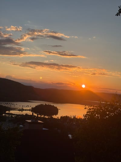 This is a vertical, eye-level, outdoor, full shot photograph of a sunset over a body of water, likely Lac Tremblant, in Mont-Tremblant, Canada. The sun is a bright, orange orb positioned just above the horizon, casting a warm glow over the landscape. Wispy, golden-orange clouds are scattered across the sky, contrasting with the clear blue sky above. Silhouetted mountains form the backdrop, their dark forms softened by the evening light. In the foreground, a body of water reflects the sunset's colors, with a small island dotted with trees in the middle. On the shore of the lake, there are visible boats, as well as buildings and a parking lot. The image is taken from an elevated vantage point, looking down on the scene, with dark trees in the lower right corner framing the view. The overall impression is one of tranquility and natural beauty.