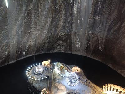 This image shows an interior view of the Salina Turda salt mine in Turda, Romania. The vast cavernous space is characterized by its layered, striated salt walls, which appear dark gray and brown with lighter white streaks, suggesting geological formations.
In the center of the image, a man-made attraction is situated on a rocky platform overlooking a dark body of water. This attraction features a circular viewing platform with a radially arranged, illuminated railing, resembling a sun or a wheel. Adjacent to this is a futuristic, capsule-shaped structure made of glass and metal, with illuminated lights. Further along, a large, spherical sculpture constructed from a metallic frame and illuminated bulbs sits on the platform. Wooden walkways and staircases connect these elements, allowing visitors to navigate the area.
Several people are visible on the platform, walking and observing the attractions, suggesting it is a tourist destination. The lighting from the attractions creates a warm, inviting atmosphere, contrasting with the dark, natural setting of the salt mine. The time of day is indeterminate due to the underground location, but the artificial lighting implies it is open for visitors. The overall mood is one of awe and wonder at the unique combination of natural geological beauty and human-made entertainment. There is no visible text in the image.
