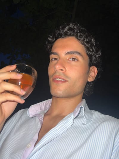A young man with curly dark hair, wearing a light blue and white striped button-down shirt with a pink collar, holds a glass of amber liquid in his left hand. He is looking at the camera, with his mouth slightly open. The background is dark, with some shadowy foliage visible. The image is taken at night, possibly outdoors. The location is Saint-Nicolas, Belgium.