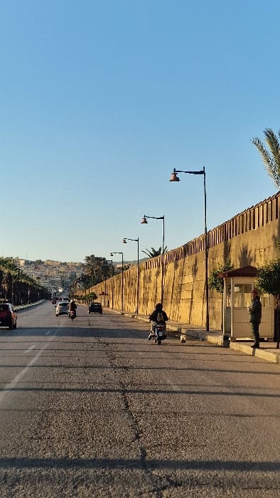 The image presents a wide-angle view of a multi-lane asphalt road in Mechouar Fes Jdid, Morocco, under a clear, light blue sky, illuminated by sunlight casting long shadows from right to left.
On the right, a tall, light-colored retaining wall, possibly stone or concrete, runs parallel to the road, featuring a patterned top edge. A concrete sidewalk borders the wall, along which multiple black street light poles with orange-hued, bell-shaped lamps are installed. A small, white rectangular guard booth is situated on this sidewalk, near the foreground. A lone individual, dressed in a khaki/green uniform and a red cap, stands to the right of the guard booth, facing left towards the road. Palm trees are visible behind and above the retaining wall.
The asphalt road exhibits visible cracks and surface wear. Multiple vehicles are in motion, proceeding away from the viewer. These include several passenger cars, such as a red sedan in the left foreground and a silver car in the mid-ground, along with at least two individuals riding motorcycles or scooters in separate lanes.
The left side of the road is lined with palm trees and other green foliage. In the distant background, a densely built hillside city or town is discernible beneath the clear sky.