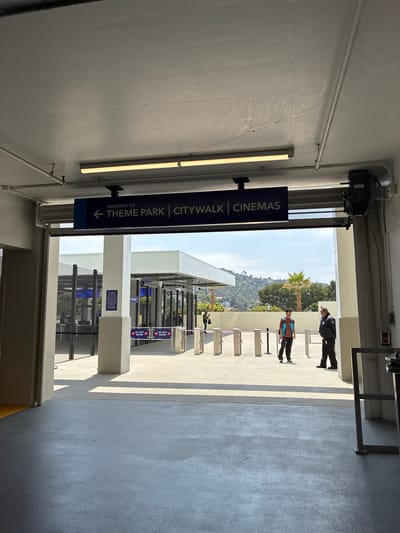 An exterior, eye-level, medium shot shows an entrance to a pedestrian walkway that leads to a theme park and cinemas. The walkway is covered by a concrete overhang with fluorescent lighting. A dark blue sign with white text hangs from the overhang, reading "WALKWAY TO ← THEME PARK | CITYWALK | CINEMAS."
Beyond the entrance, the walkway opens to a sunlit area with multiple ticket turnstiles. Two men are visible near the turnstiles. One, wearing a dark jacket and pants, appears to be a security guard. The other, a younger man in a teal vest over an orange shirt and black pants, stands facing the security guard. Behind the turnstiles, a tall fence encloses an area with more structures and trees. In the background, a lush green hillside rises under a clear blue sky with scattered clouds. The environment suggests a warm, sunny day in a location with a mild climate. The overall scene depicts an access point to a public entertainment venue.