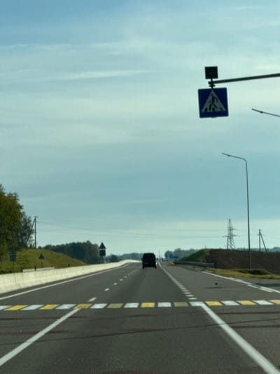 The image depicts a multi-lane asphalt highway on a partially overcast day in Пяршайскі сельскі Савет, Belarus. The road, viewed from a moving vehicle, extends into the distance, with a dark-colored van or large car visible ahead in the right lane, receding from the viewer. In the foreground, a pedestrian crosswalk is marked across the width of the road by alternating yellow and white rectangular blocks. Overhead, a blue square sign with a white pedestrian symbol indicates the crossing.
The highway features solid white lines delineating the outer edges and dashed white and yellow lines for lane separation. To the left, a concrete barrier separates the road from a grassy embankment topped with deciduous trees. Further along the left side, a dark, triangular road sign is visible, along with utility poles and power lines. On the right, a tall, lattice-style electricity pylon stands on a raised earthen area. A street light pole with a single luminaire extends over the right shoulder. The distant horizon shows a faint tree line under a light blue sky with scattered clouds. No individuals are visible.