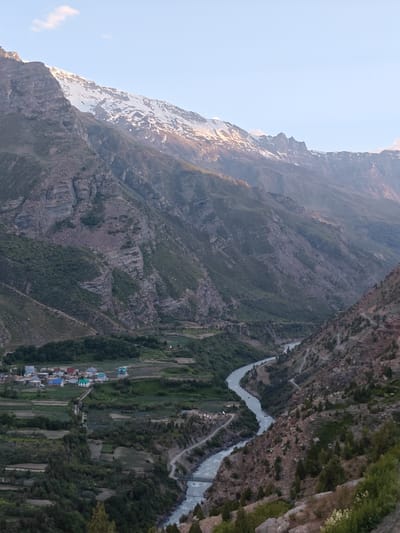 A vast, mountainous landscape under a clear, pale blue sky. The scene is dominated by towering, rugged mountains, some of which are capped with snow. The slopes are steep and rocky, with patches of green vegetation clinging to them. A river snakes through the valley below, its waters a pale, milky blue.
In the valley, a small village is nestled amongst terraced fields and scattered trees. The houses are a mix of colors, with many featuring bright blue or red roofs. A road winds its way through the landscape, connecting the village to the wider area. The time of day appears to be late afternoon or early evening, as the light casts long shadows and highlights the textures of the mountains. The weather is clear, with a single wisp of cloud in the sky. The overall mood is one of serene, natural beauty and peaceful isolation.
There is no visible text in this image. The location is identified as Gemur, India.