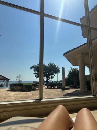 The image presents an indoor-to-outdoor view through a multi-pane window, captured from a low, reclining perspective. In the immediate foreground, the bare, tanned thighs and knees of an individual are visible, partially obscuring a light-colored surface. To the left of the legs, a small portion of a transparent glass object is visible. Below the windowpanes, a light-colored sill or baseboard runs horizontally, featuring a two-receptacle electrical outlet on its left side.
Beyond the window, a bright, sunlit exterior is depicted under a clear blue sky, exhibiting a distinct lens flare in the upper right quadrant, indicating direct sunlight. A large body of water, consistent with a lake, stretches across the mid-ground, with a calm surface. The immediate outdoor area is a spacious patio paved with irregular flagstones. Prominent vegetation includes manicured hedges, a large mature tree with a broad, dark green canopy centered in the mid-ground, and additional smaller trees and dense foliage to the right. On the left side of the water, a covered boat slip with a reddish-brown tiled roof shelters a boat. On the patio, to the right of the central tree, an outdoor dining set consisting of a rectangular table and multiple chairs is arranged. Further right, a large, un-erected patio umbrella base with a central pole is visible. The right side of the outdoor scene reveals a section of a large building, likely a residence, featuring a red-tiled roof, light-colored walls, and substantial classical columns.
This scene is consistent with a waterfront residential property located on Lake Conroe, near Montgomery, Texas, United States.