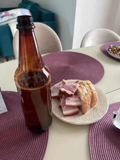 The image shows a table setting, possibly indoors. A partially full brown beer bottle lies horizontally across a purple placemat. Next to the bottle is a white plate containing sliced bread and what appears to be slices of bacon. There is a small dish containing nuts in the top left corner of the frame. A portion of two chairs are visible, one beige and one teal, and they appear to be in a residential setting. The location mentioned is in Хлябово, Russia, but the image does not provide enough data to verify or confirm this information.