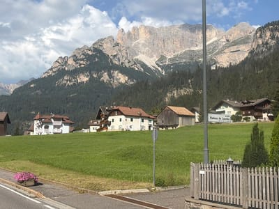 In Mazzin - Mazin, Italy, a scenic view captures a village nestled against a majestic mountain range. The foreground features a paved road with a flower box filled with pink blossoms. A weathered wooden fence with a lamppost stands to the right. Behind the fence, a lush green meadow slopes upwards, dotted with several buildings.
On the meadow, a white building with a red-tiled roof and pink balconies is prominent. To its left, another building with a white facade and a red roof is visible. Further to the right, a rustic wooden barn-like structure stands. In the background, a larger white building with a red roof displays a sign that reads "strada Madona de Lourdes".
Dominating the upper portion of the image is a towering mountain range, its rugged peaks and slopes covered in dense evergreen forest. The sky above is a mix of blue with scattered white clouds. The scene depicts a tranquil, mountainous village setting.