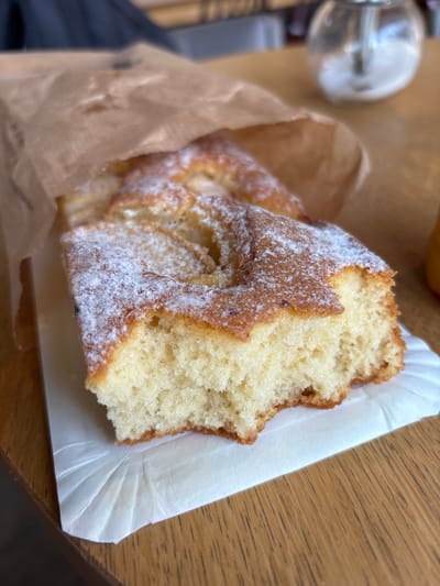 This is a close-up, eye-level photograph of a slice of cake, dusted with powdered sugar, resting on a white paper liner. The cake is partially visible, emerging from a brown paper bag. The cake itself appears to be a light, airy sponge cake with a slightly browned crust and a tender, crumbly interior. The powdered sugar is unevenly distributed, with some areas having a thicker coating than others.
The cake is positioned on a light brown, wooden surface, likely a table, which has a prominent wood grain texture. The lighting suggests it is daytime, possibly indoors given the soft illumination. The background is out of focus, but hints of a metallic object, perhaps a sugar dispenser or a part of a coffee machine, are visible to the left. The overall scene conveys a casual, perhaps a cafe or bakery setting. There is no visible text in the image.