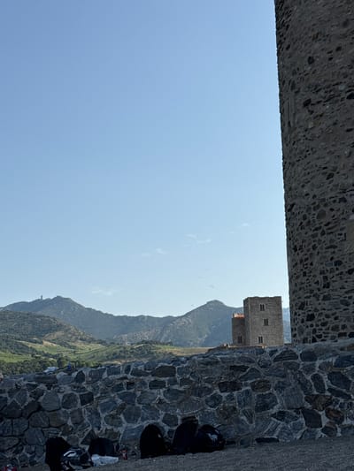 The image shows a section of a stone tower on the right, a stone wall in the foreground, and in the background, two stone towers of a building, a mountainside with vegetation, and a clear blue sky. The scene appears to be outdoors on a sunny day. The stone tower on the right is large and textured, made of various sizes of dark stones. The stone wall in the foreground is also constructed of dark, irregular stones.

In the mid-ground, two rectangular stone towers are visible. They appear to be part of a historic building, possibly a fortress or castle, with arched windows. Behind these towers, a sloping mountainside covered in green vegetation, including what looks like vineyards, is depicted. A distant mountain range is visible further in the background. A tall, thin structure, possibly a lighthouse or antenna, is perched on one of the distant mountain peaks.

Several dark backpacks are placed on the ground in front of the stone wall in the foreground. Some white items, possibly clothing or bags, are also visible near the backpacks, and a red and white bottle is also present. One backpack on the left has a white and black motorcycle helmet next to it. The ground beneath the backpacks appears to be gravel.

This scene is likely the Royal Castle of Collioure (Château Royal de Collioure), a historic fortress located on the coast of Collioure, France. The architecture of the towers and the surrounding mountainous landscape are characteristic of this location.