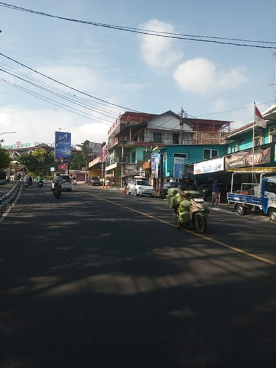 The image depicts a street scene in what appears to be an urban area. The main subject is a person riding a motorcycle heavily laden with what looks like sacks of produce, moving along a road marked with a yellow line. Other vehicles, including cars and smaller trucks, are visible on the street. Several people are seen standing near the storefronts on the right side of the image. Buildings line the street, featuring a mix of architectural styles and colors. A banner advertising a product is prominently displayed. Overhead, power lines crisscross the sky, partially obscured by some clouds. A flag with red and white stripes is visible, and signage indicates the presence of a "SPKLU PLN ELECTRIC VEHICLE CHARGING STATION KELISTRIKAN." The image suggests a busy commercial area with a mix of traffic and pedestrian activity. The location is potentially in the city of Zeyathiri District, Myanmar.