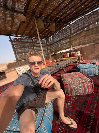 The image shows a young man with curly blonde hair and sunglasses sitting on a blue cushion in an outdoor seating area. He is wearing a grey t-shirt and brown shorts. He is looking towards the camera and holding his right hand up. The seating area is covered with a thatched roof and decorated with woven baskets hanging from the ceiling. There are several large, patterned cushions on the floor, and a wooden table with benches in the background. The overall setting appears to be a desert environment, consistent with Agafay, Morocco, known for its desert landscapes and traditional accommodations. The lighting suggests it is daytime.