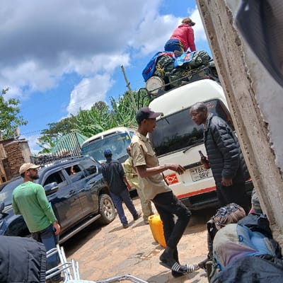 The scene depicts a busy outdoor area in Mweka, Tanzania, under a partly cloudy blue sky. In the immediate foreground on the right, a rough, light-colored concrete or stone wall partially obscures silver metal chairs and dark backpacks on the ground. The ground throughout the visible area is paved with uneven, light-colored stones or bricks.

In the midground, two men are interacting. The man on the left, wearing a beige short-sleeved collared shirt, black pants, black socks, and black-and-white slide sandals, with a black baseball cap, is gesturing with his left hand. The man on the right, wearing a black puffer jacket and dark trousers, holds a brown glass bottle in his right hand. A yellow jerry can rests on the ground near the feet of the man in the beige shirt.

Behind these men, a white van (Tanzanian license plate "T869 DRV") is stationary. Its roof rack is heavily laden with cargo, including bundles covered by blue and green tarps, and several white, round containers. A person wearing a pink long-sleeved top and blue jeans is seated on top of this luggage, facing away from the camera.

To the left of the white van, a dark-colored SUV is parked. A bearded man in a green long-sleeved shirt and dark pants stands next to the SUV's driver's side, looking towards the left. An arm and head are partially visible inside the SUV through the rear passenger window. Further left and slightly behind the SUV, a man in a dark shirt and pants walks away from the camera. Another person in light-colored attire is partially visible between the SUV and the white van.

The background features dense green vegetation, including what appear to be trees, and a utility pole. A portion of another white and yellow bus is visible behind the black SUV. On the far left, a brick building structure is present.