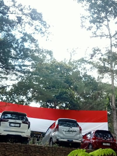 The image depicts an outdoor scene in Kuwukan, Indonesia. In the foreground, a low retaining wall constructed of irregular brown and grey stones is visible, with green foliage on its right side. Immediately behind this wall, three vehicles are parked facing away from the observer. From left to right, these are: a white Toyota Innova with license plate B 1924 KZJ, a light silver Toyota Avanza with license plate B 2367 KB, and a red compact hatchback with license plate B 1788 PI. Directly behind the vehicles, a large horizontal banner displaying the red (upper) and white (lower) colors of the Indonesian flag is prominently displayed. Partially visible behind this banner is a dark structure with the word "coffee" written in white letters, indicating a business establishment. The upper half of the frame is dominated by a bright, possibly overcast sky and a dense canopy of green trees. No individuals are present.