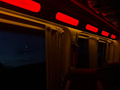 The interior of a train car at night. Red rectangular lights illuminate the ceiling. Tan curtains hang over the windows. Outside the window, a dark landscape is visible. The background is completely black. Seats are in the foreground. The location of Anipemza, Armenia, is not discernible from the image.
