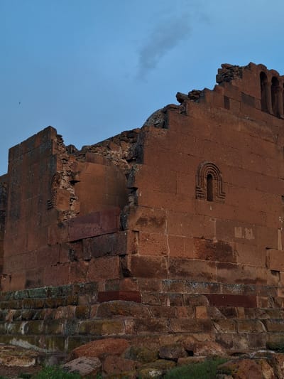 The image shows a section of a ruined building in Anipemza, Armenia. The building is constructed of red brick. The wall is in a state of disrepair, with significant portions of the brickwork missing. There is a decorative carving resembling a "U" shape. The background shows a blue sky.