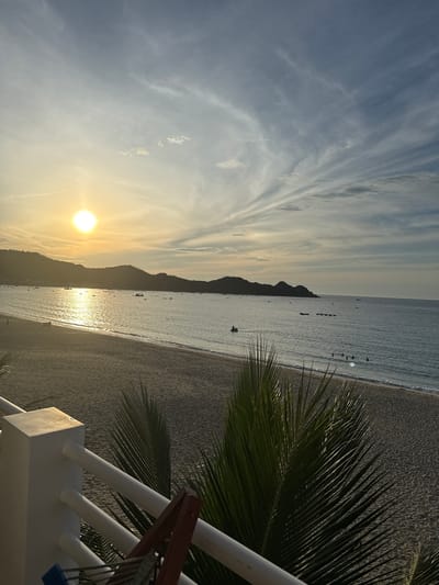 This is a photograph taken from a raised vantage point, likely a balcony or railing, overlooking a beach and the sea at sunset in Song Cau, Vietnam. The sun, a bright orb, is setting on the left side of the frame, casting a golden glow across the calm ocean and reflecting off the water in a shimmering path. In the distance, a dark, hilly coastline stretches across the horizon, silhouetted against the sky. Several small boats are scattered across the water, some closer to shore and others further out. On the beach, the sand appears light brown and gently slopes towards the water. A few figures are visible on the beach and in the shallows of the sea, suggesting recreational activity. In the foreground, the fronds of a palm tree partially obscure the view, adding a tropical element to the scene. The sky is a soft gradient of blue and white, with wispy clouds streaked across it. The overall impression is one of tranquility and the end of a day.
