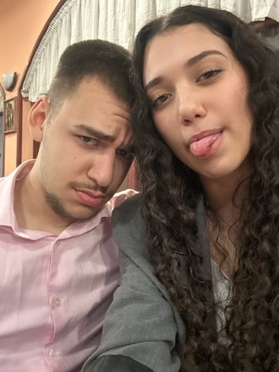 An indoor, close-up photograph depicts two young adults. On the left, a male with short dark hair, light stubble, and a pink collared shirt is shown from the chest up, looking directly at the camera with a slight frown and pursed lips. To his right, a female with long, dark, curly hair, wearing a gray blazer over a light-colored top, is also looking directly at the camera, with her tongue extended in a playful gesture. Their heads are close together. The background on the left features a light peach-colored wall with a wall-mounted light fixture and a dark-framed picture. The upper central and right background shows white, draped fabric or curtains. The exact location within Arkovna, Bulgaria, cannot be determined from the visual information provided.