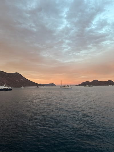 The image depicts a body of water, likely a bay or harbor, with mountains lining the shoreline in an unknown location in Mexico. The sky is split in appearance; one side is filled with clouds, and the other is a hazy, orange hue, possibly indicating either sunrise or sunset. Several boats are visible in the water near the shoreline, appearing to be docked or anchored. The water's surface is slightly rippled, suggesting a light breeze. The mountain slopes appear arid.