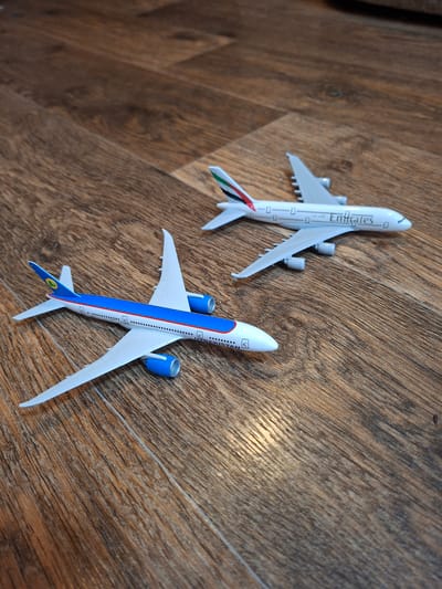 Two model airplanes are on a wooden floor. The airplane in the foreground has a blue stripe along its side and the logo of Uzbekistan Airways on its tail. The airplane in the background is white with the Emirates logo on its side. The floor is made of wooden planks with a visible wood grain.